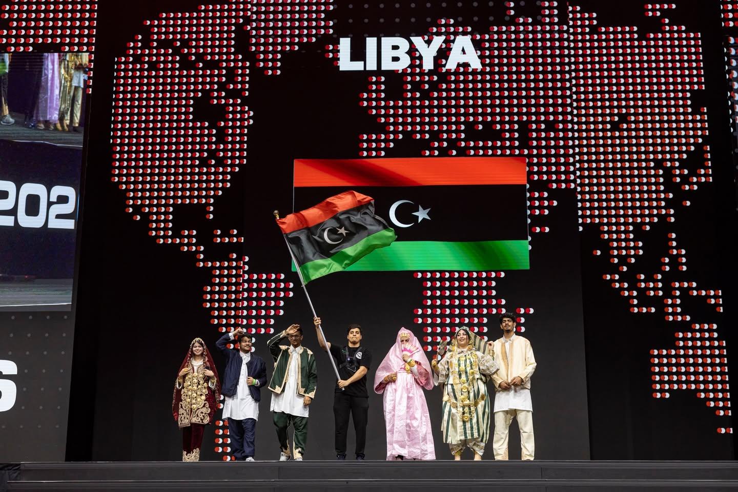 طلابنا على المسرح في بطولة العالم VEX للروبوتات في سانت لويس، أبريل 2026، ومعظمهم بالزي الليبي التقليدي، وأحدهم يلوّح بعلم ليبيا تحت شاشة كُتب عليها LIBYA