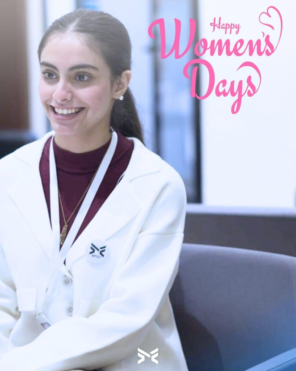 شابة مبتسمة ترتدي معطفاً أبيض وشارة MTG7، بجانب عبارة Happy Women's Days.