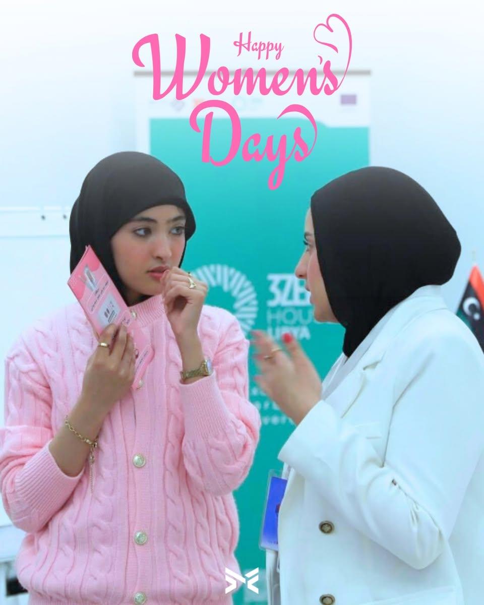 شابتان بحجاب أسود تتحدثان قرب لافتة فيروزية، وإحداهما تمسك بطاقة وردية.