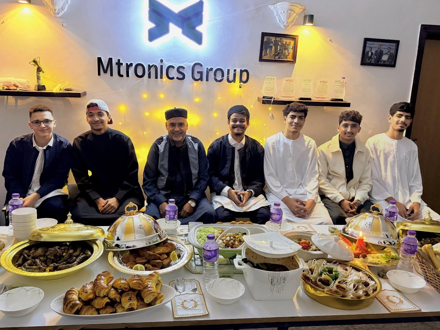 سبعة أشخاص بالزي التقليدي خلف مائدة إفطار عامرة، أسفل شعار Mtronics Group المضاء.