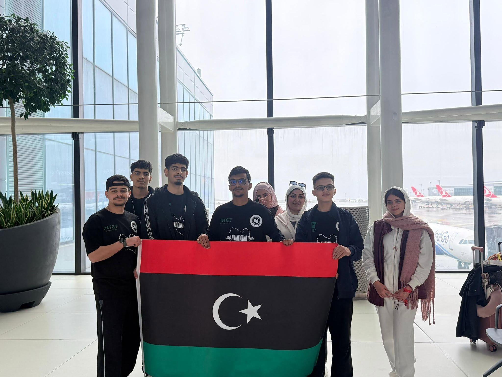ثمانية أشخاص، بعضهم بقمصان فريق MTG7، يحملون علماً ليبياً كبيراً داخل صالة مطار.