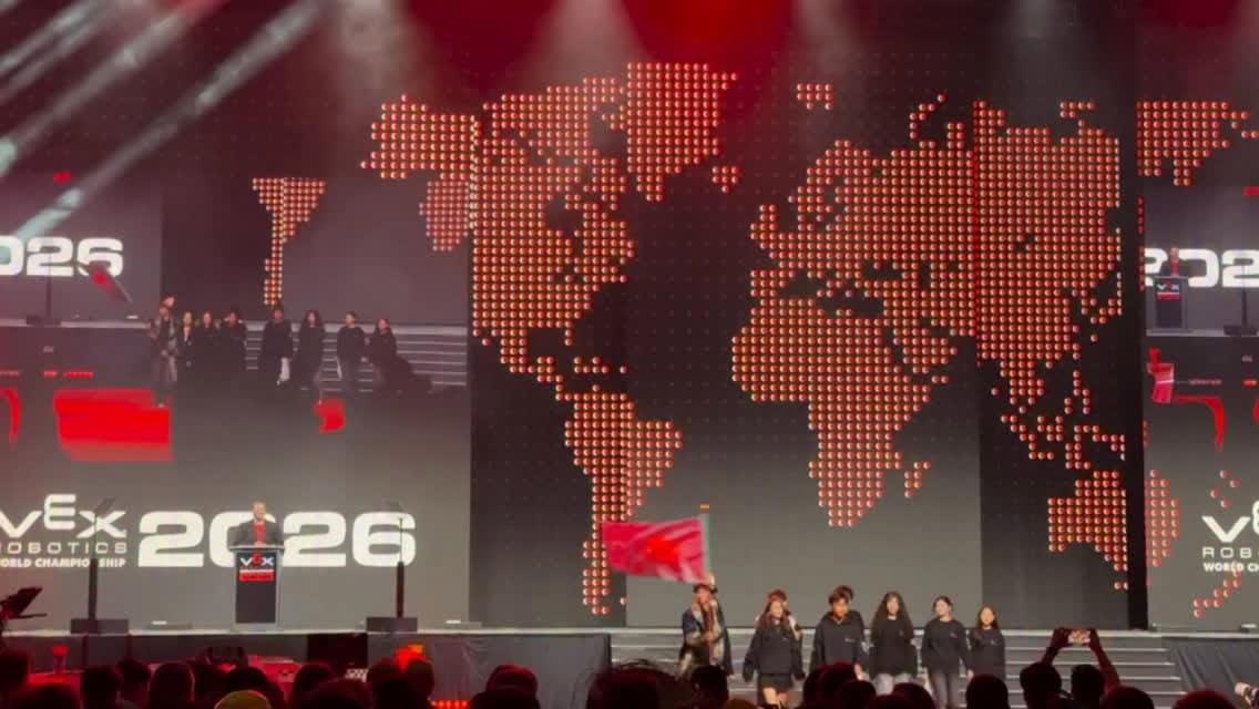منصة حفل الافتتاح مع شاشات VEX 2026 وخريطة عالم حمراء منقطة وأشخاص يسيرون حاملين راية.