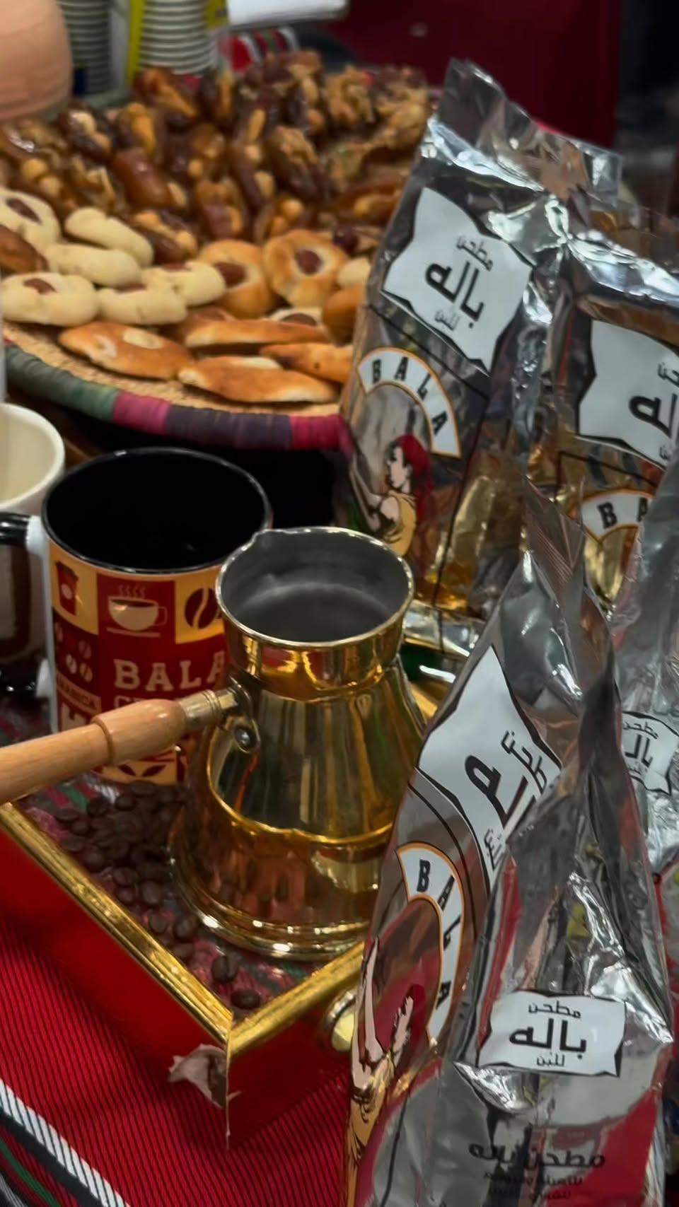 إبريق قهوة نحاسي وأكياس قهوة باله وصينية حلويات وتمر على قماش أحمر.