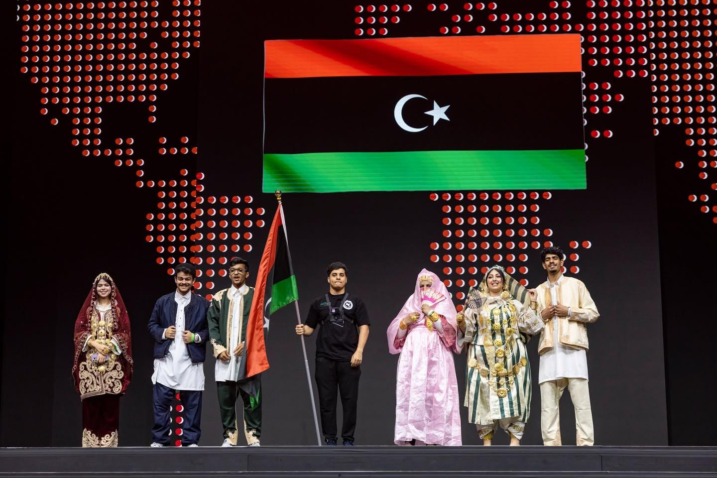 طلاب، معظمهم بالزي الليبي التقليدي، على المسرح تحت شاشة ضخمة تعرض العلم الليبي