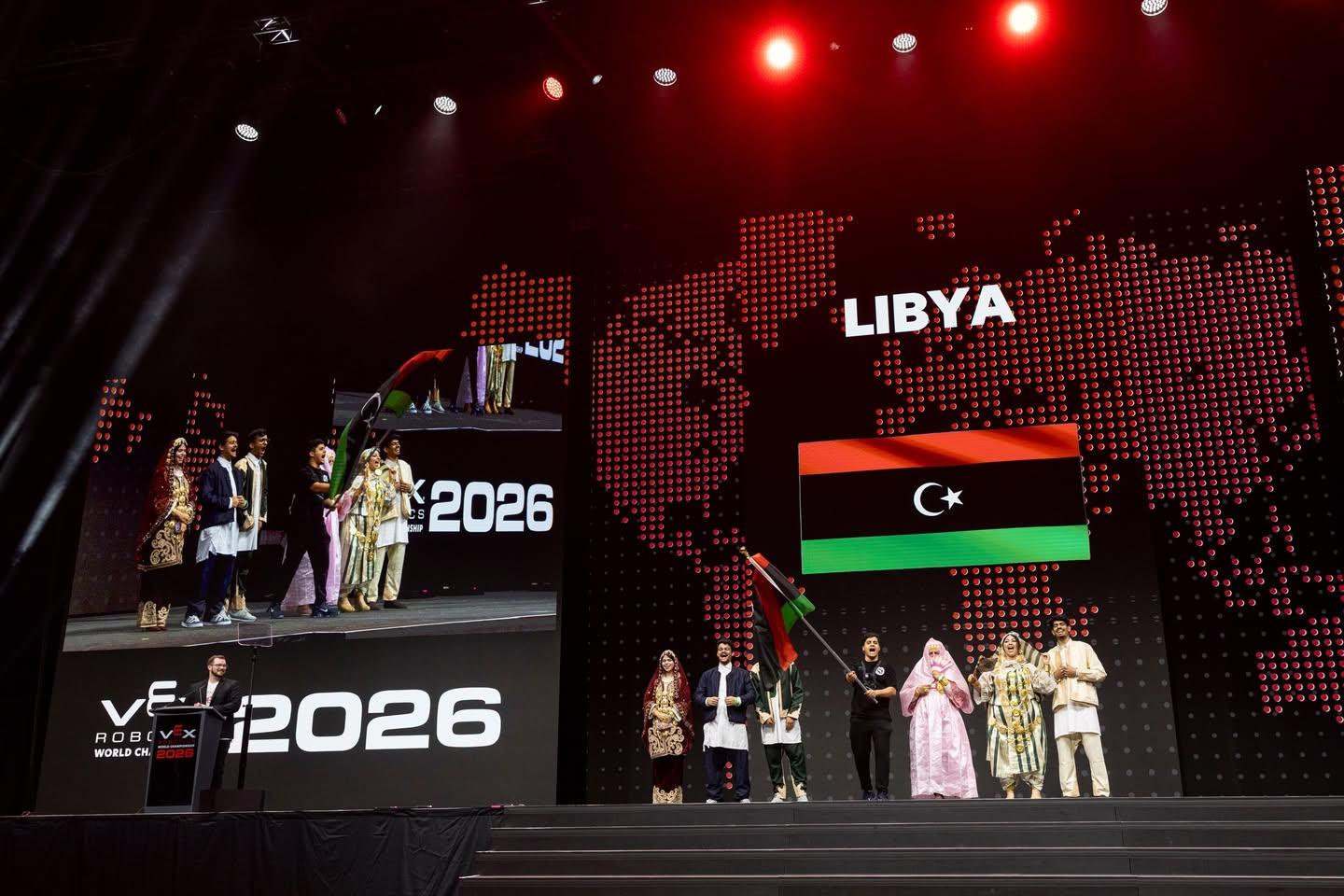 طلاب على المسرح، معظمهم بالزي الليبي التقليدي، وأحدهم يلوّح بالعلم تحت شاشة كُتب عليها LIBYA