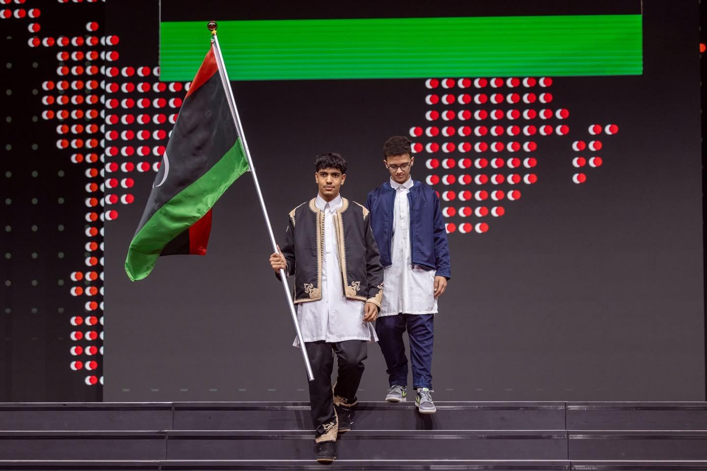 طالبان بالزي الليبي التقليدي ينزلان درجات المسرح، أحدهما يحمل العلم الليبي