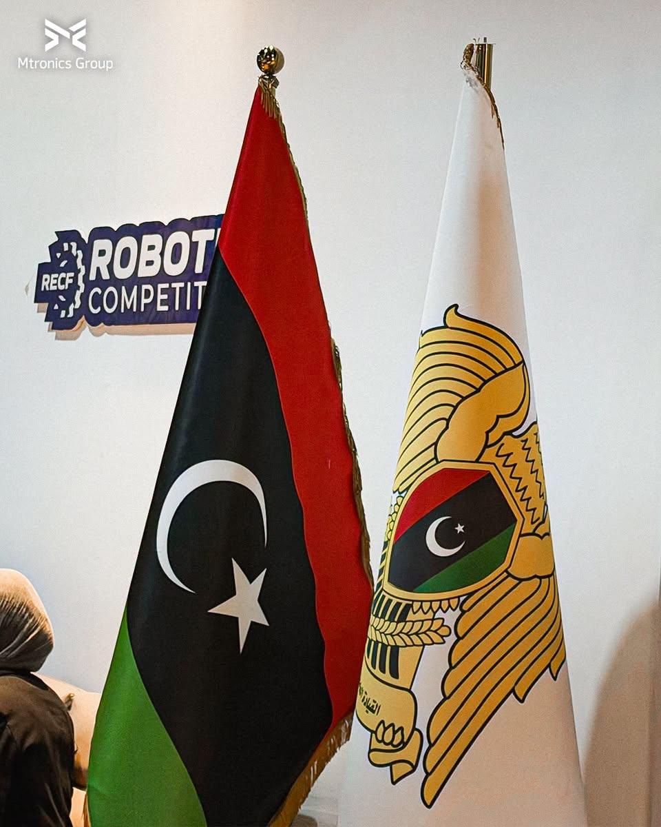 العلم الليبي وعلم القيادة العامة على حوامل، وخلفهما لافتة مسابقة الروبوتات من RECF